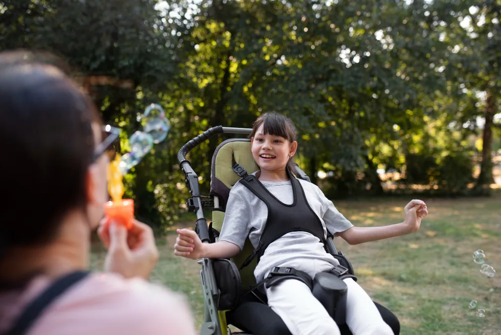A girl in a supportive wheelchair laughing at soap bubbles blown by a carer outdoors
